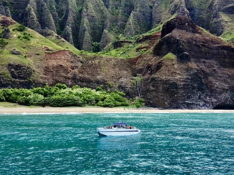 A small boat off a beach below green cliffs and a thin waterfall
