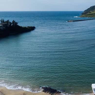 A calm bay with a sandy beach, a low wooded point and a breakwater