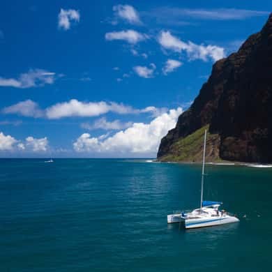 A catamaran at rest in deep blue water below a dark headland