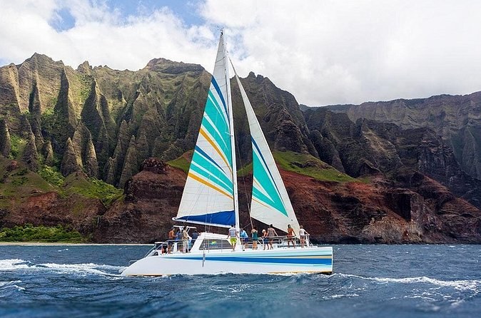 A catamaran under striped sails running along steep ridged cliffs