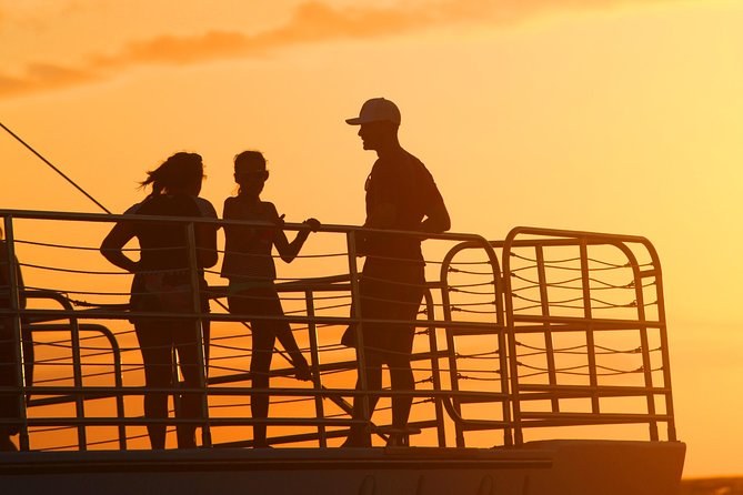 Passengers in silhouette at a boat's rail against an orange sunset sky